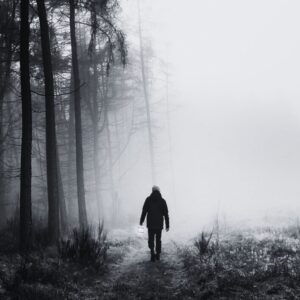 A black and white image of the back of a shadowy male looking individual on a path holding a lantern sluggishly at waist height. To the right, there is part of a clearing. To the left there is a very dense forest with lots of tall trees.
